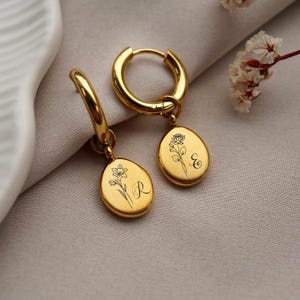 May include: Gold hoop earrings with oval pendants. Each pendant has a floral design and a letter, one with an "R" and the other with an "E". The earrings are presented on a neutral-toned surface.