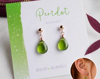 May include: A pair of drop earrings featuring teardrop-shaped green peridot stones suspended from rose gold-toned posts. The earrings are displayed on a white card printed with the text Peridot, August, and Growth Relaxation.