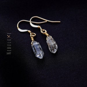 Puede incluir: Un par de pendientes de alambre dorado envueltos con gotas de cristal de cuarzo transparente. Los pendientes están sobre un fondo negro.