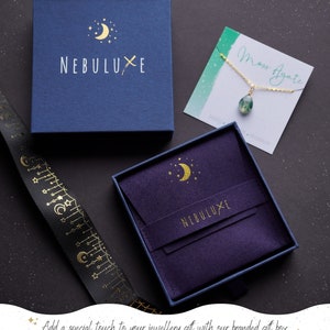 May include: A purple velvet jewelry box with a gold crescent moon and stars design. The box is open and has a blue outer box with the text "NEBULUXE" in gold lettering. The box is perfect for storing and gifting jewelry.