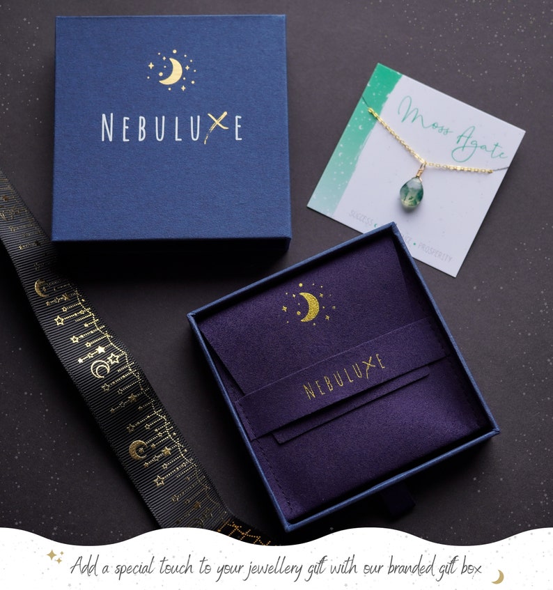 May include: A purple velvet jewelry box with a gold crescent moon and stars design. The box is open and has a blue outer box with the text "NEBULUXE" in gold lettering. The box is perfect for storing and gifting jewelry.