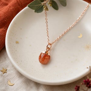 Può includere: Una collana in oro rosa con un ciondolo in pietra del sole arancione, di forma ovale e sfaccettata. La catenina sottile è fissata a una capsula metallica che trattiene la gemma. La collana è appoggiata in un piccolo piattino in ceramica color crema.