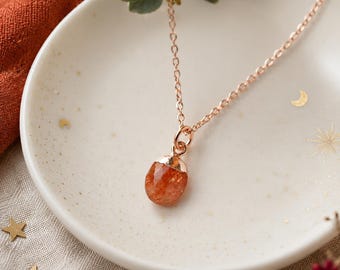May include: A rose gold necklace featuring a faceted, oval-shaped orange sunstone pendant. The delicate chain is attached to a metal cap securing the gemstone. The necklace is displayed inside a small, off-white ceramic dish with gold star and moon accents.
