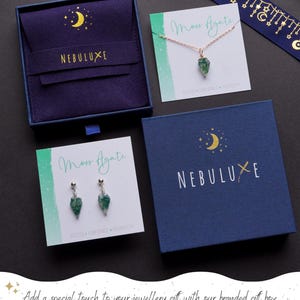 May include: An assortment of jewelry and gift boxes. The image features a necklace and earrings with green stone pendants, displayed on cards that read "Moss Agate." The boxes are navy blue with gold crescent moon and star designs, and the brand name "NEBULUXE."