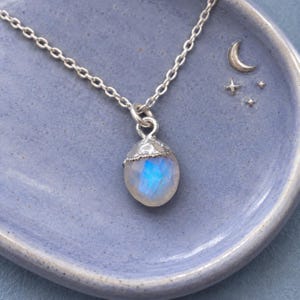 May include: A silver chain necklace featuring a faceted, teardrop-shaped moonstone pendant with a textured silver cap. The stone displays a blue iridescent glow, and the pendant is suspended from a delicate, polished silver link chain.