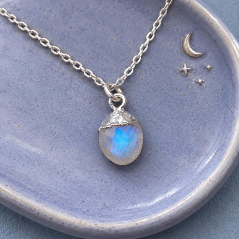 May include: A silver chain necklace featuring a faceted, teardrop-shaped moonstone pendant with a textured silver cap. The stone displays a blue iridescent glow, and the pendant is suspended from a delicate, polished silver link chain.