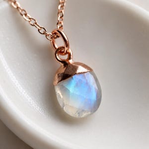 May include: A rose gold necklace featuring a faceted, teardrop-shaped moonstone pendant. The stone displays a translucent white appearance with iridescent blue flashes. The pendant is secured by a rose gold cap and hangs from a delicate chain.