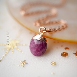 Op de afbeelding: Een roségouden ketting met een gefacetteerde, dieproze edelsteen hanger. De hanger is bedekt met goud en hangt aan een delicate ketting. Het woord "NEBULUXE" staat verticaal aan de linkerkant van de afbeelding.