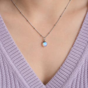 May include: A silver-toned necklace featuring a delicate chain with small evenly spaced metal beads and a single, small, teardrop-shaped iridescent stone pendant that displays subtle blue and white light reflections against a metallic setting.