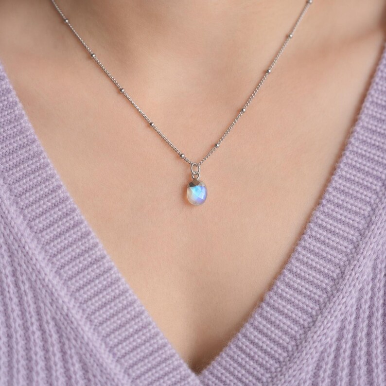 May include: A silver-toned necklace featuring a delicate chain with small evenly spaced metal beads and a single, small, teardrop-shaped iridescent stone pendant that displays subtle blue and white light reflections against a metallic setting.
