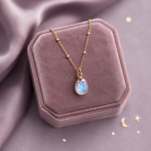 May include: A gold chain necklace featuring a delicate satellite chain design and a single teardrop-shaped moonstone pendant with a faceted surface. The pendant displays a soft blue iridescent glow, suspended from a simple gold metal bail.
