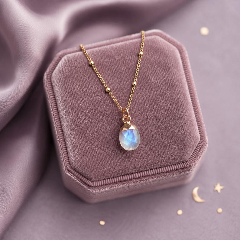 May include: A gold chain necklace featuring a delicate satellite chain design and a single teardrop-shaped moonstone pendant with a faceted surface. The pendant displays a soft blue iridescent glow, suspended from a simple gold metal bail.