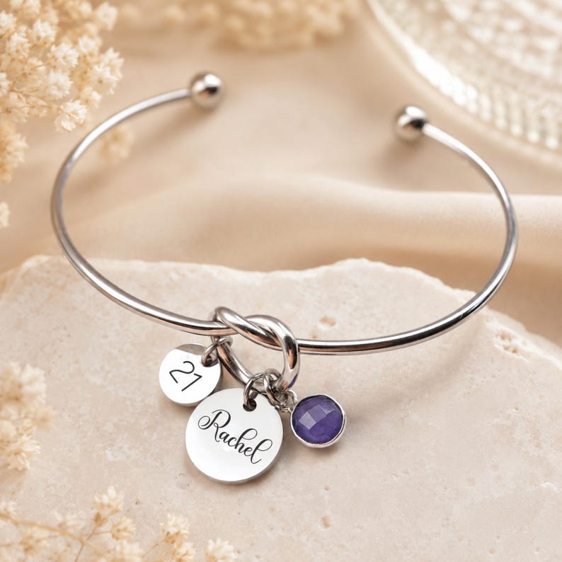 May include: A silver-toned metal cuff bracelet featuring a central knot design. Three charms hang from the knot: a small circular disc engraved with the number 21, a larger circular disc engraved with the name Rachel, and a faceted purple stone.