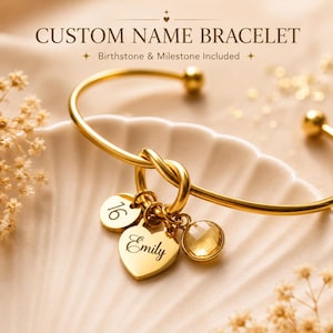 May include: A gold-toned metal bangle bracelet featuring a central knot design. Three charms are attached: a round disc engraved with the number 16, a heart-shaped charm engraved with the name Emily, and a round, faceted yellow birthstone charm.
