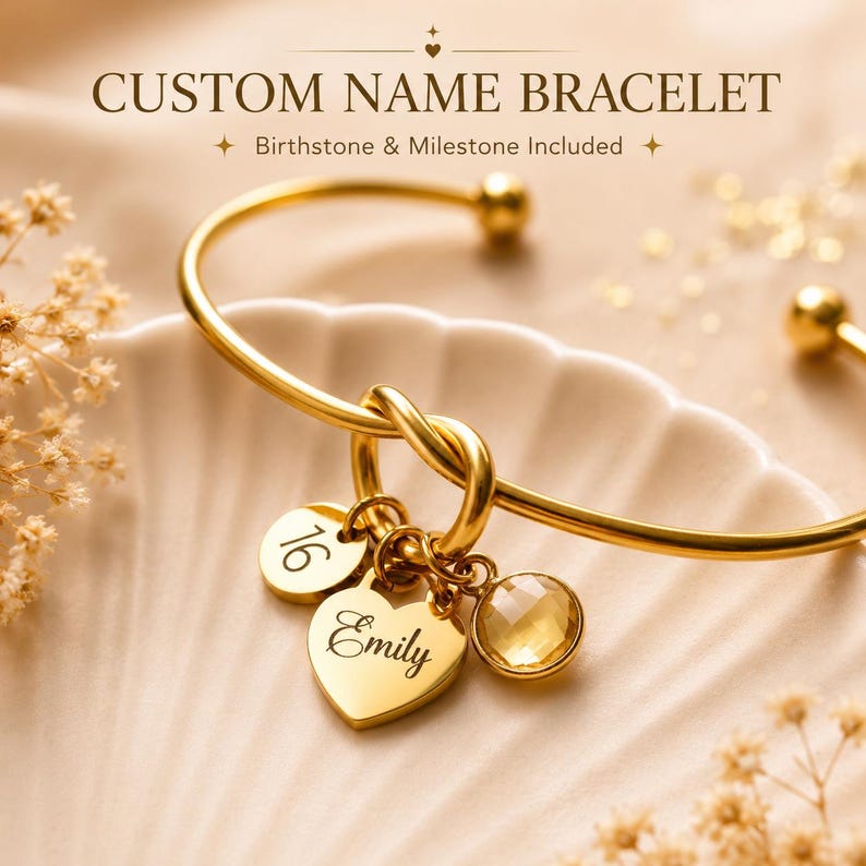 May include: A gold-toned metal bangle bracelet featuring a central knot design. Three charms are attached: a round disc engraved with the number 16, a heart-shaped charm engraved with the name Emily, and a round, faceted yellow birthstone charm.