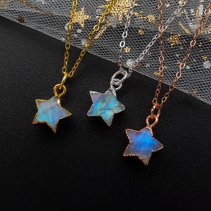 以下が含まれることがあります： 虹色の青い星型のペンダントが付いた、シルバー、ゴールド、ローズゴールドのネックレス3本。ネックレスは黒い背景の上に置かれています。