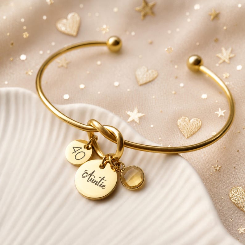 May include: A gold-toned cuff bracelet featuring a knot detail and three dangling charms. One charm is engraved with the number 40, another with the word Auntie in script, and the third is a faceted, round, light-colored gemstone charm.