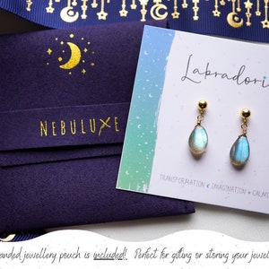 May include: A pair of gold labradorite earrings with a teardrop shape. The earrings are displayed on a card with the text "Labradorite" and "Transformation Imagination Calming". A blue velvet pouch with the text "NEBULUXE" is included.