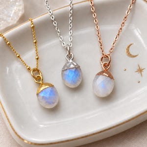 May include: Three necklaces featuring faceted, oval-shaped moonstone pendants with blue iridescence. Each pendant is capped in metal and suspended from a delicate chain. The metals are gold, silver, and rose gold, matching the chain and cap colours.