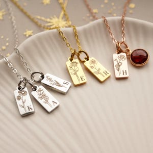 May include: Four silver necklaces with delicate floral charms and initial tags. The charms are engraved with the letters K, A, S, and E. The necklace on the right is rose gold with a red gemstone and the letter H.