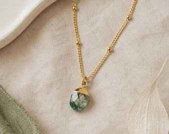 May include: A gold chain necklace featuring a delicate satellite bead design and a single oval-shaped moss agate pendant. The pendant is capped with a smooth gold metal setting, showcasing the natural green and white inclusions of the stone.