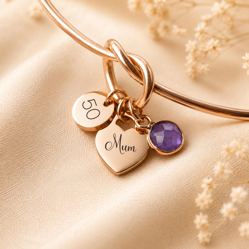 May include: A rose gold bangle bracelet featuring a knot detail and three charms: a circular disc engraved with the number 50, a heart-shaped charm engraved with the word Mum in script, and a small round faceted purple gemstone charm.