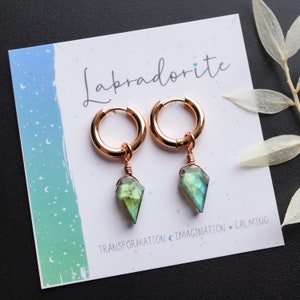 May include: Rose gold hoop earrings with faceted labradorite pendants. The earrings are displayed on a white card with the word "Labradorite" and the words "Transformation, Imagination, Calming".