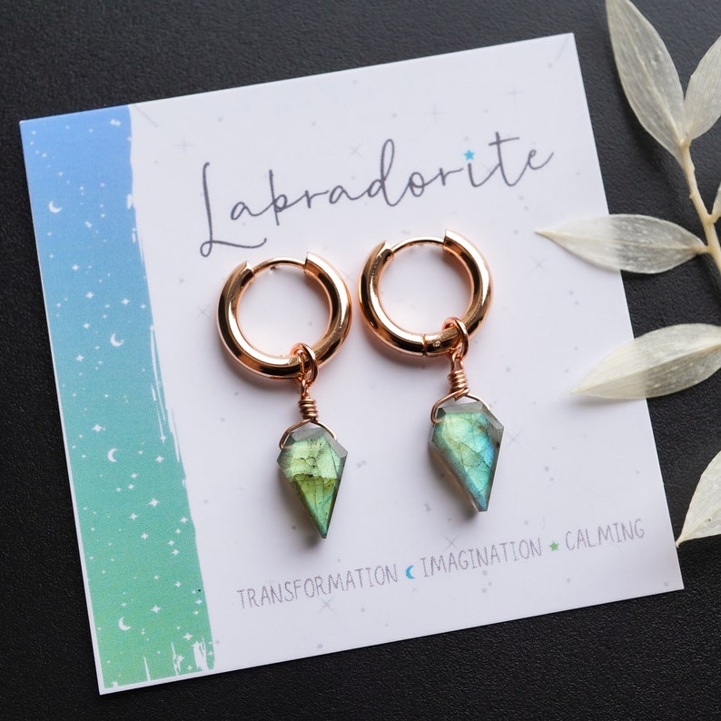 May include: Rose gold hoop earrings with faceted labradorite pendants. The earrings are displayed on a white card with the word "Labradorite" and the words "Transformation, Imagination, Calming".