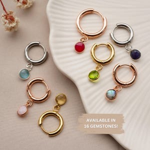 Peut inclure: Plusieurs boucles d'oreilles créoles en or, or rose et argent, chacune ornée d'une pierre précieuse différente. Les boucles d'oreilles sont présentées sur un plat blanc festonné. Le texte "AVAILABLE IN 16 GEMSTONES!" est visible.