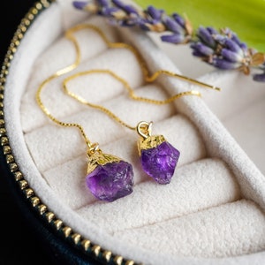Puede incluir: Un par de pendientes de oro con gotas de piedras preciosas de amatista púrpura. Los pendientes se muestran en una caja de joyería de terciopelo.