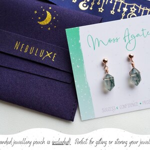 May include: A pair of moss agate earrings with rose gold wire wrapped around the stones. The earrings are on a white card with the text "Moss Agate" and "SUCCESS CONFIDENCE PROSPERITY". The earrings are packaged in a blue velvet pouch with a gold crescent moon and stars design and the text "NEBULUXE".