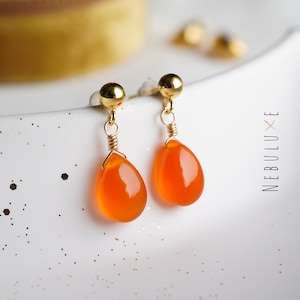 May include: A pair of gold-toned earrings with orange teardrop-shaped stones. The earrings are made with wire wrapped around the stones.