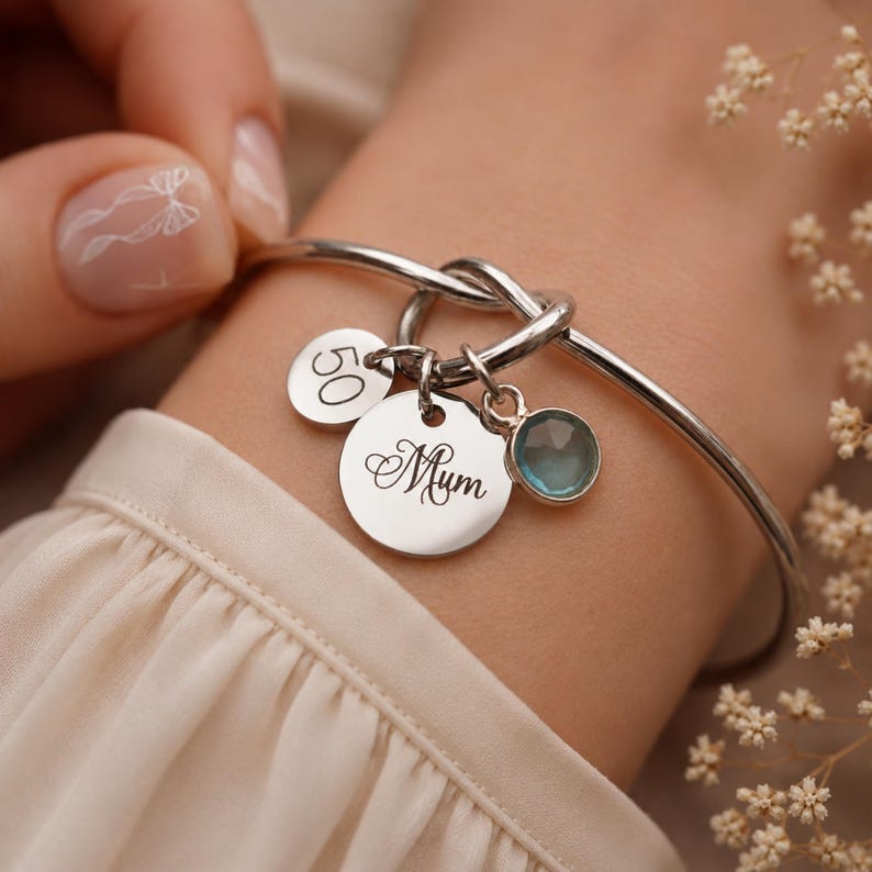 May include: A silver-toned bangle bracelet featuring a knot detail and three charms: a small circular disc engraved with the number 50, a larger circular disc engraved with the word Mum in script, and a round, faceted light blue gemstone charm.