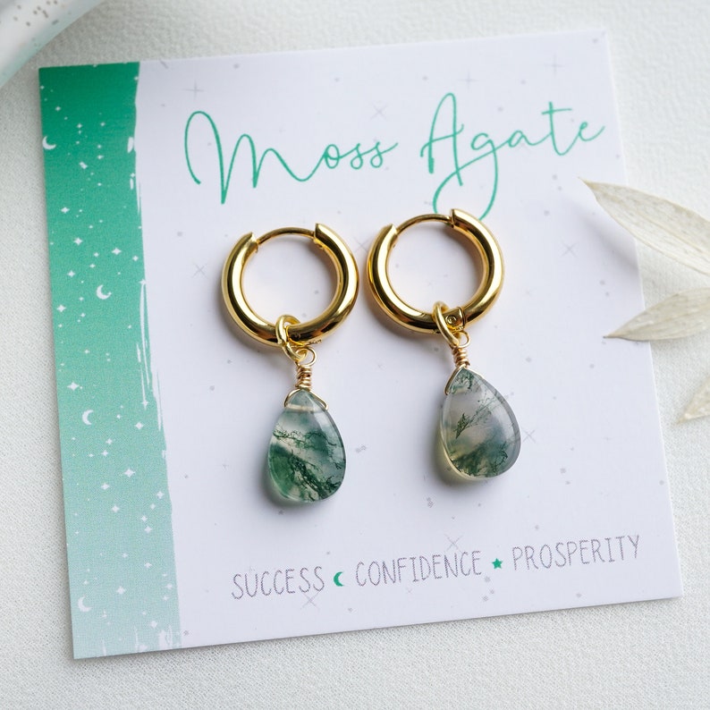 May include: Gold hoop earrings with moss agate teardrop charms. The earrings are displayed on a white card with the text "Moss Agate", "Success", "Confidence", and "Prosperity".
