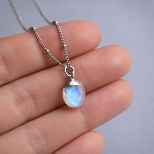 May include: A silver necklace featuring a delicate chain with small metal beads and a faceted, teardrop-shaped moonstone pendant. The stone displays a translucent white appearance with a distinct blue iridescent glow set in a silver cap.