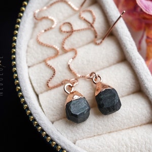 Op de afbeelding: Een paar zwarte edelsteen oorbellen met roségouden accenten en een delicate ketting. De oorbellen worden gepresenteerd in een witte fluwelen doos.