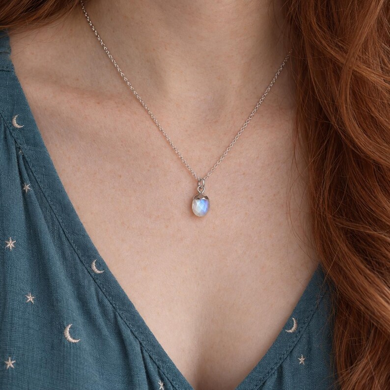 May include: A silver chain necklace featuring a small, oval-shaped moonstone pendant with a smooth, iridescent surface. The pendant is securely held by a simple silver cap and hangs from a delicate, thin silver link chain resting against the chest.