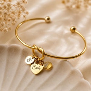 May include: A gold-toned metal cuff bracelet featuring a knot detail and three dangling charms: a circular disc engraved with the number 16, a heart-shaped charm engraved with the name Emily, and a small, round, translucent yellow gemstone charm.