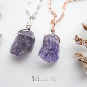 May include: Two amethyst crystal pendants on silver and rose gold chains. The crystals are raw and have a purple hue. The chains are delicate and have a simple design. The pendants are displayed on a white background with dried flowers.