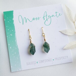 May include: A pair of moss agate earrings with gold wire wrapped around the stones. The earrings are on a white card with the text "Moss Agate" and "Success, Confidence, Prosperity".
