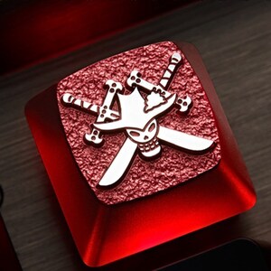May include: A red mechanical keyboard keycap with a silver skull and crossbones design. The skull is wearing a hat and the crossbones are swords.
