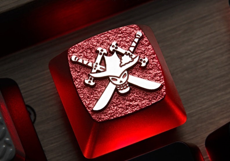 May include: A red mechanical keyboard keycap with a silver skull and crossbones design. The skull is wearing a hat and the crossbones are swords.
