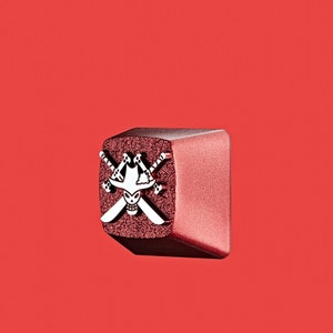 May include: A red and silver keycap with a skull and crossbones design. The skull is wearing a hat and the crossbones are swords.