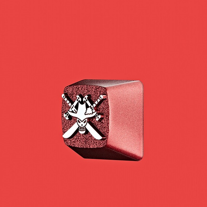May include: A red and silver keycap with a skull and crossbones design. The skull is wearing a hat and the crossbones are swords.
