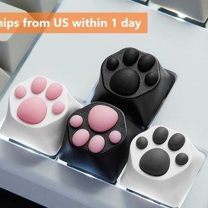 May include: Four different cat paw-shaped keycaps for a keyboard. Two keycaps are black with pink paw prints, one is white with pink paw prints, and one is white with gray paw prints.