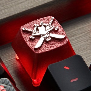 May include: A red mechanical keyboard keycap with a silver skull and crossbones design. The keycap is illuminated from below, casting a red glow.