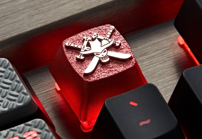 May include: A red mechanical keyboard keycap with a silver skull and crossbones design. The keycap is illuminated from below, casting a red glow.
