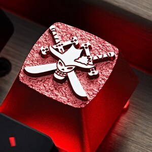 May include: A red mechanical keyboard keycap with a white skull and crossbones design. The skull is wearing a straw hat and the crossbones are swords.