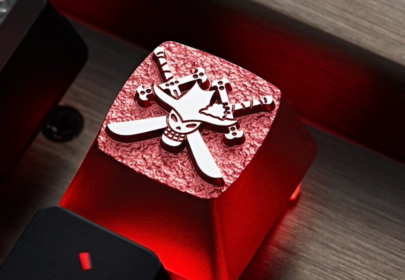 May include: A red mechanical keyboard keycap with a white skull and crossbones design. The skull is wearing a straw hat and the crossbones are swords.