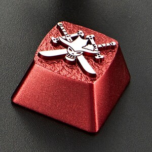May include: A red metal keycap with a silver skull and crossbones design. The skull is wearing a hat and the crossbones are crossed swords.